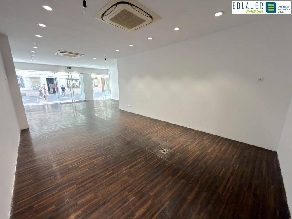 Heller Geschäftsraum mit dunklem Holzfußboden, weißen Wänden und Blick auf die belebte Straße durch die Glasfront.