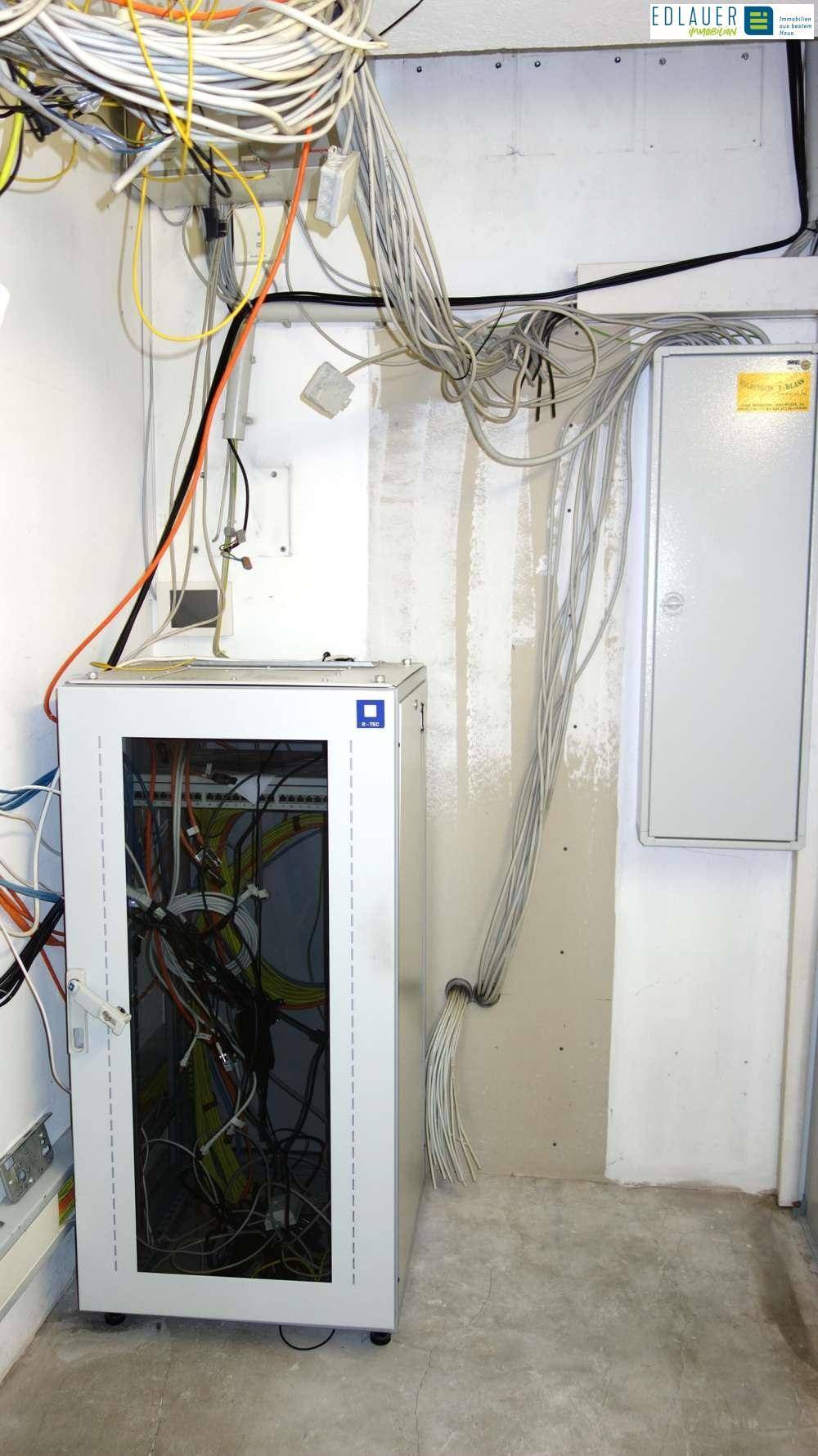 Serverraum mit einem Serverschrank, vielen Kabeln und einem Sicherungskasten an der Wand für die Netzwerkverteilung.