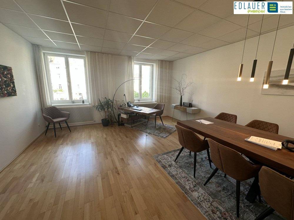 Heller Büroraum mit Holzboden, großem Besprechungstisch und zwei Fenstern mit Tageslicht.