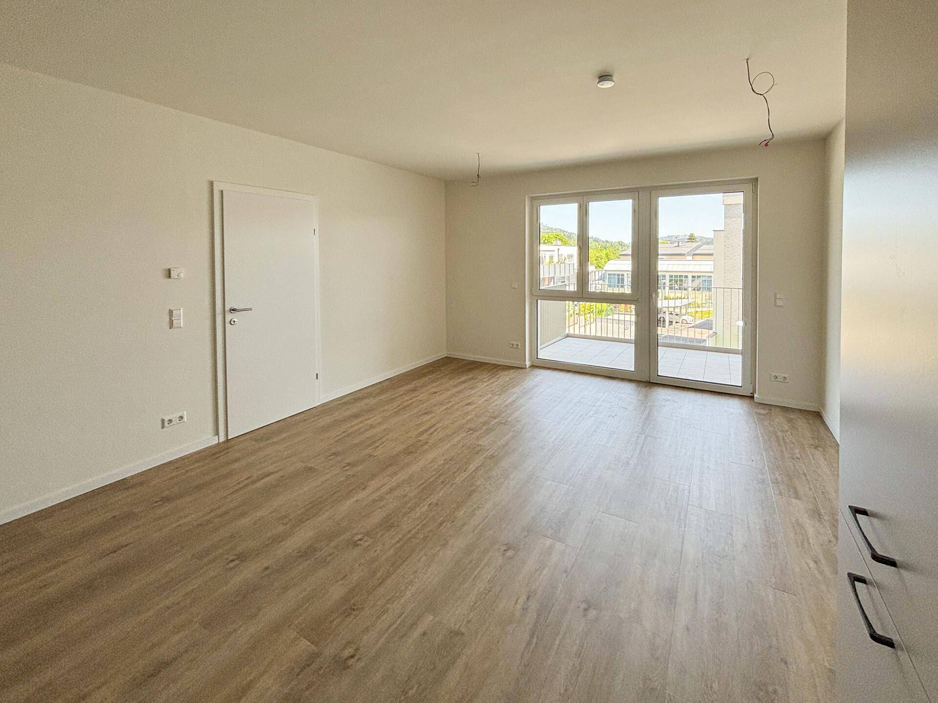 Heller, leerer Wohnraum mit Laminatboden und großen Fenstern, die Zugang zum Balkon bieten.