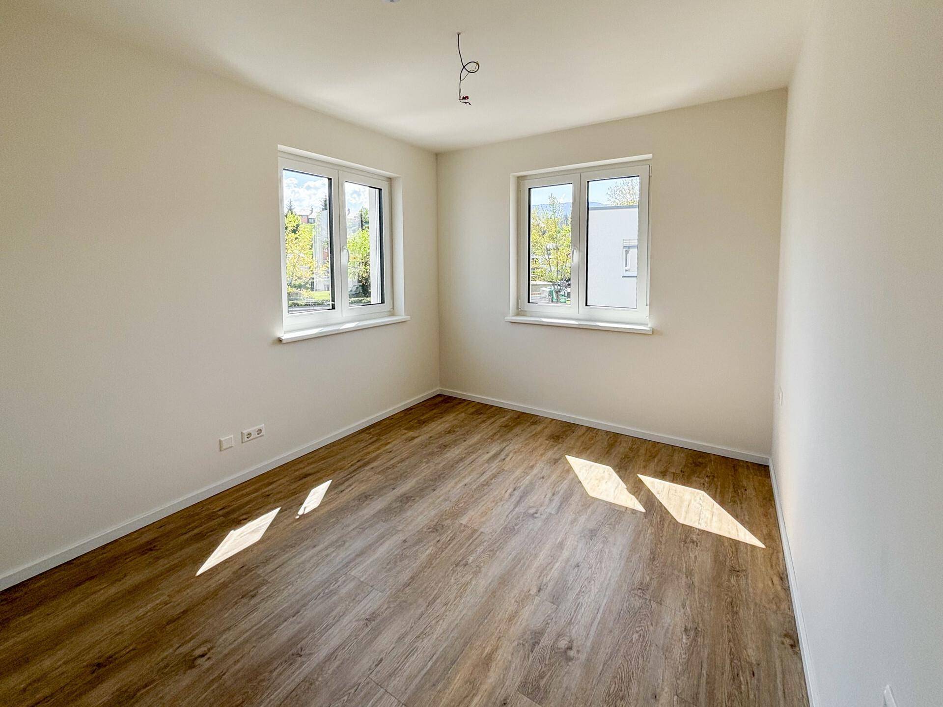 Leeres Schlafzimmer mit Laminatboden und zwei Fenstern, die viel Tageslicht hereinlassen.