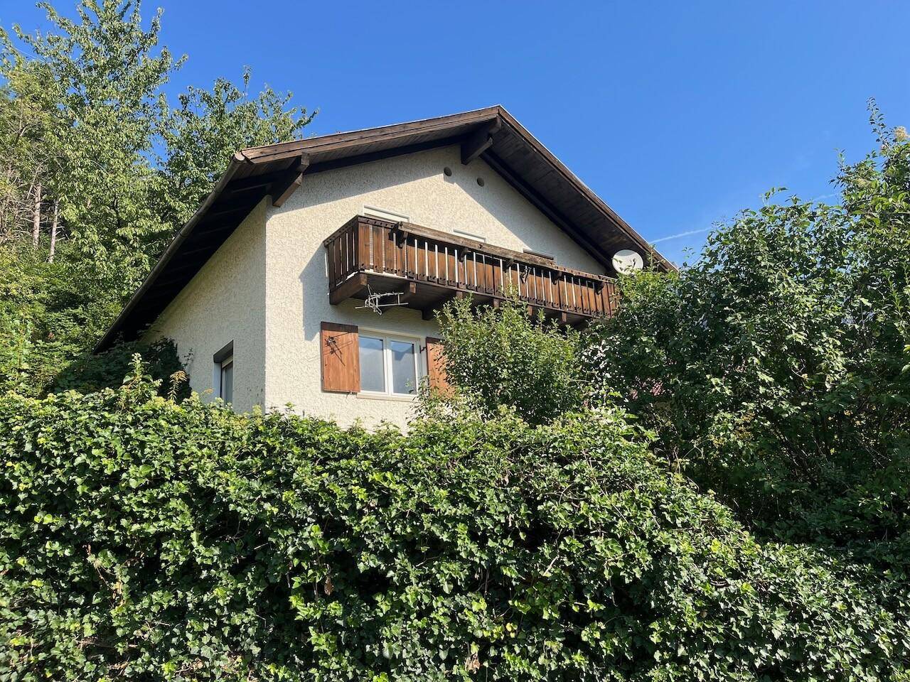 Hausfassade mit Holzbalkon und grüner Hecke, umgeben von üppiger Vegetation.