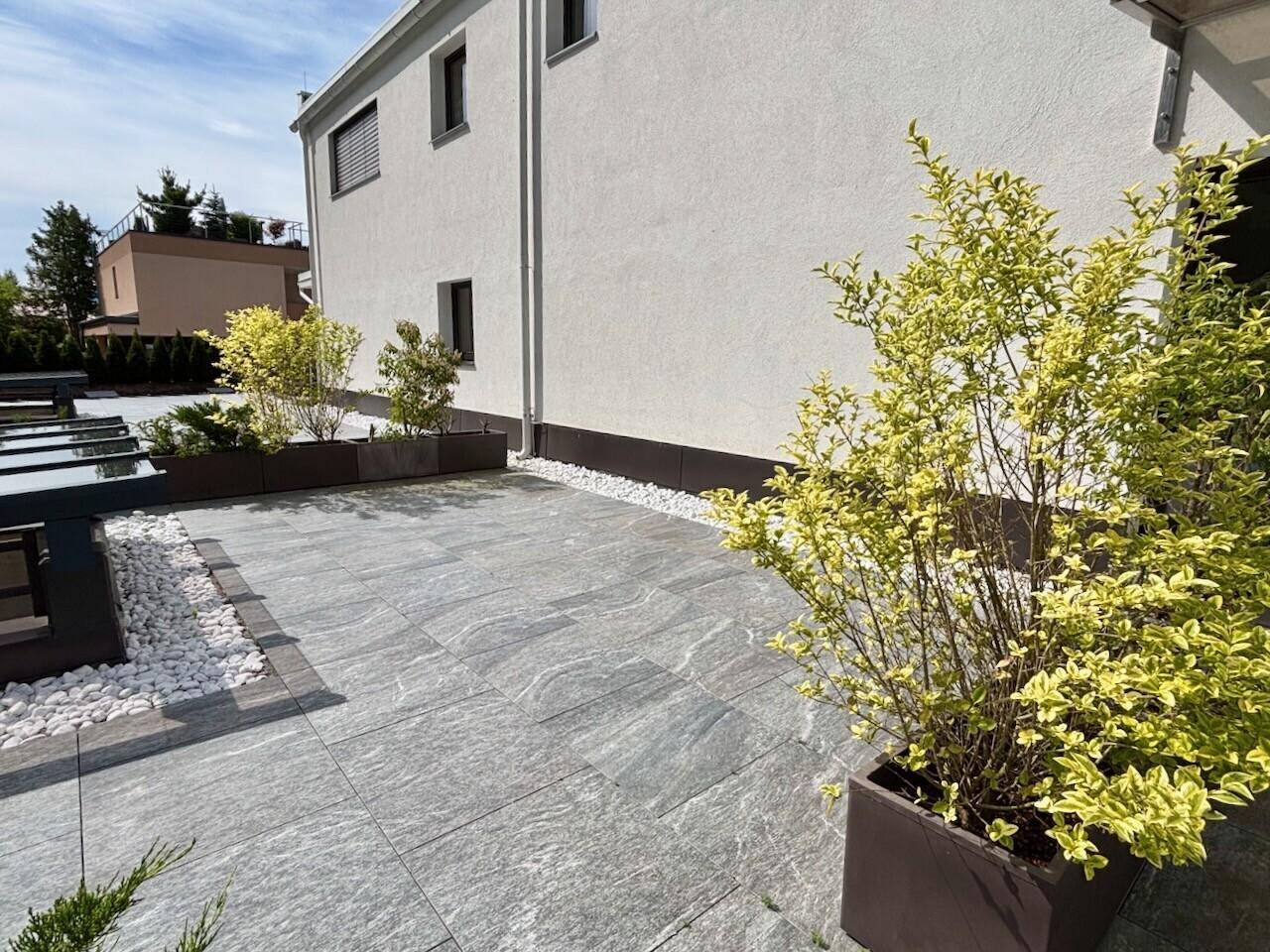 Gepflasterte Terrasse mit dekorativen Pflanzenkübeln und Kiesstreifen neben dem Gebäude.