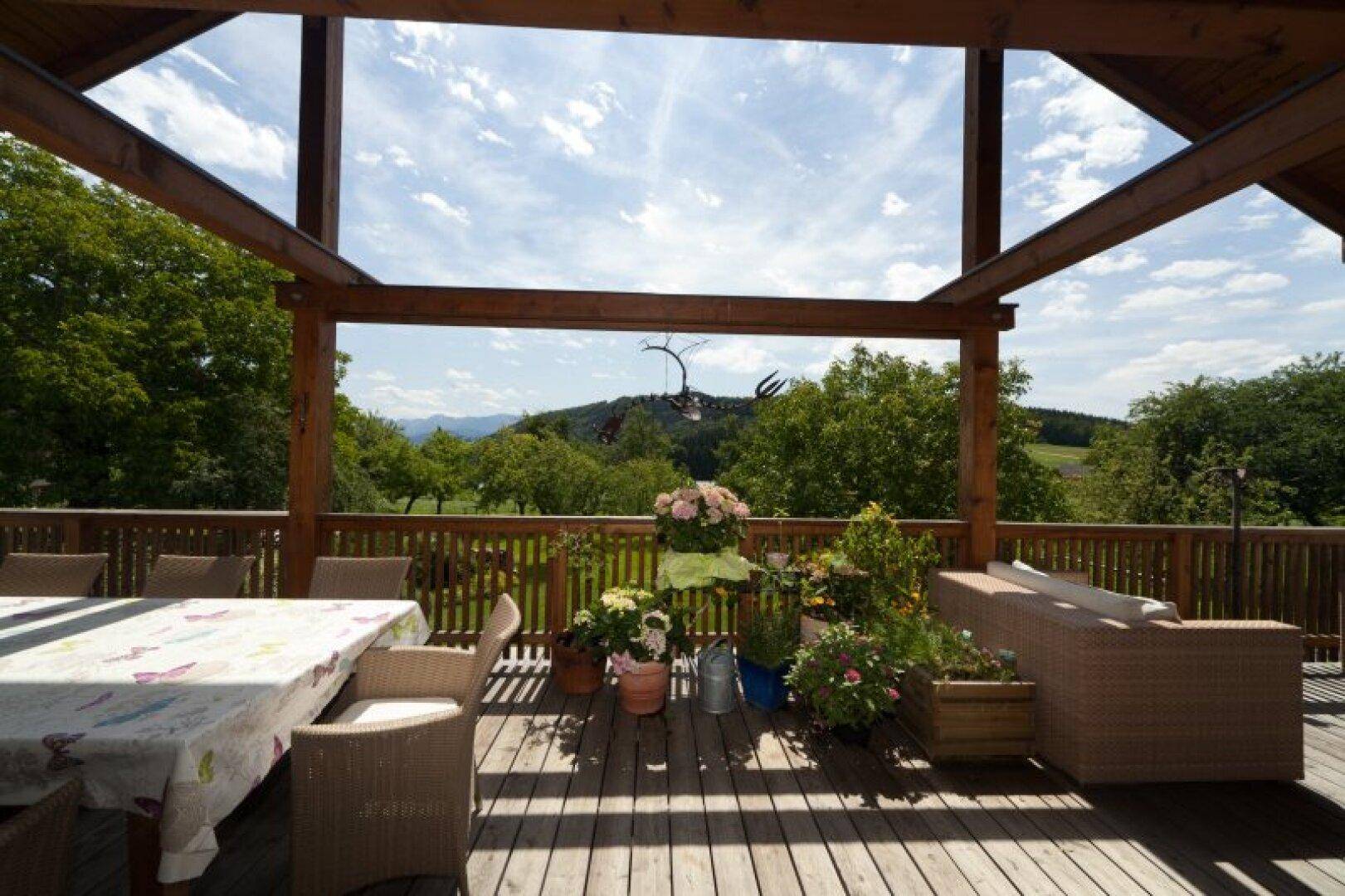 Große Holzterrasse mit Esstisch und Lounge-Bereich, umgeben von grüner Natur und Bergblick.
