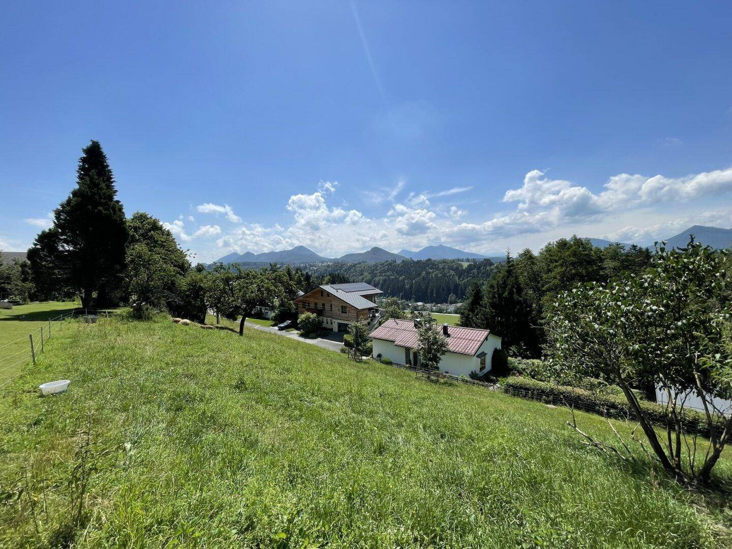Weitläufiger Blick über grüne Wiesen und Häuser auf eine malerische Berglandschaft unter blauem Himmel.