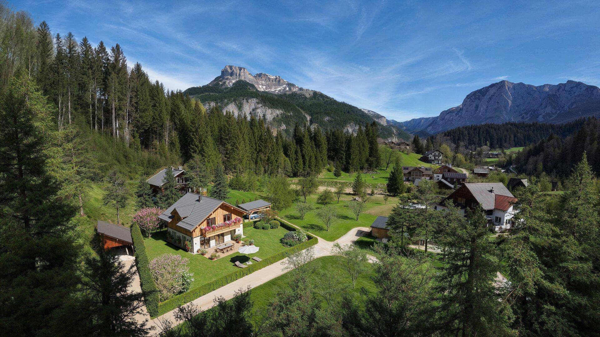 Luftaufnahme eines Hauses mit großem Garten, umgeben von Wäldern und Bergen in einer ländlichen Umgebung.