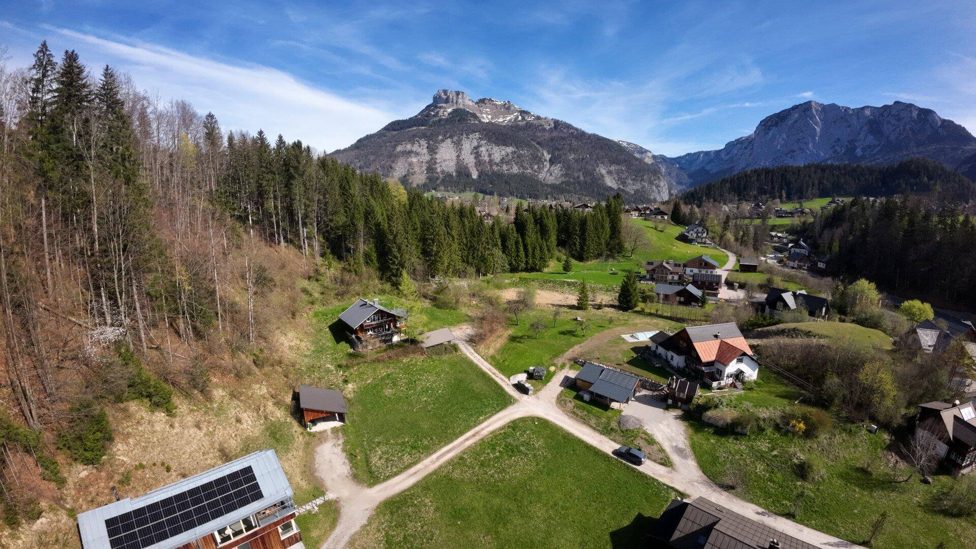 Weitläufige Luftaufnahme einer ländlichen Siedlung mit Häusern, Gärten und Solaranlagen vor einer Bergkulisse.
