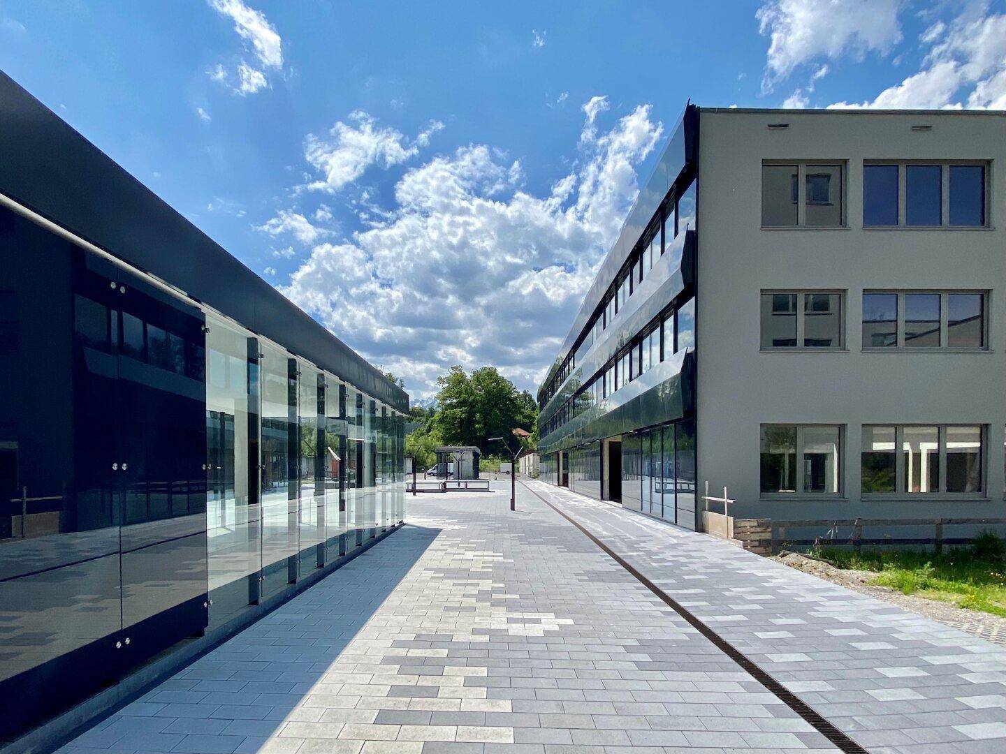 Moderne Gebäudefassaden mit Glas und grauen Elementen, umgeben von einem gepflasterten Weg unter blauem Himmel.