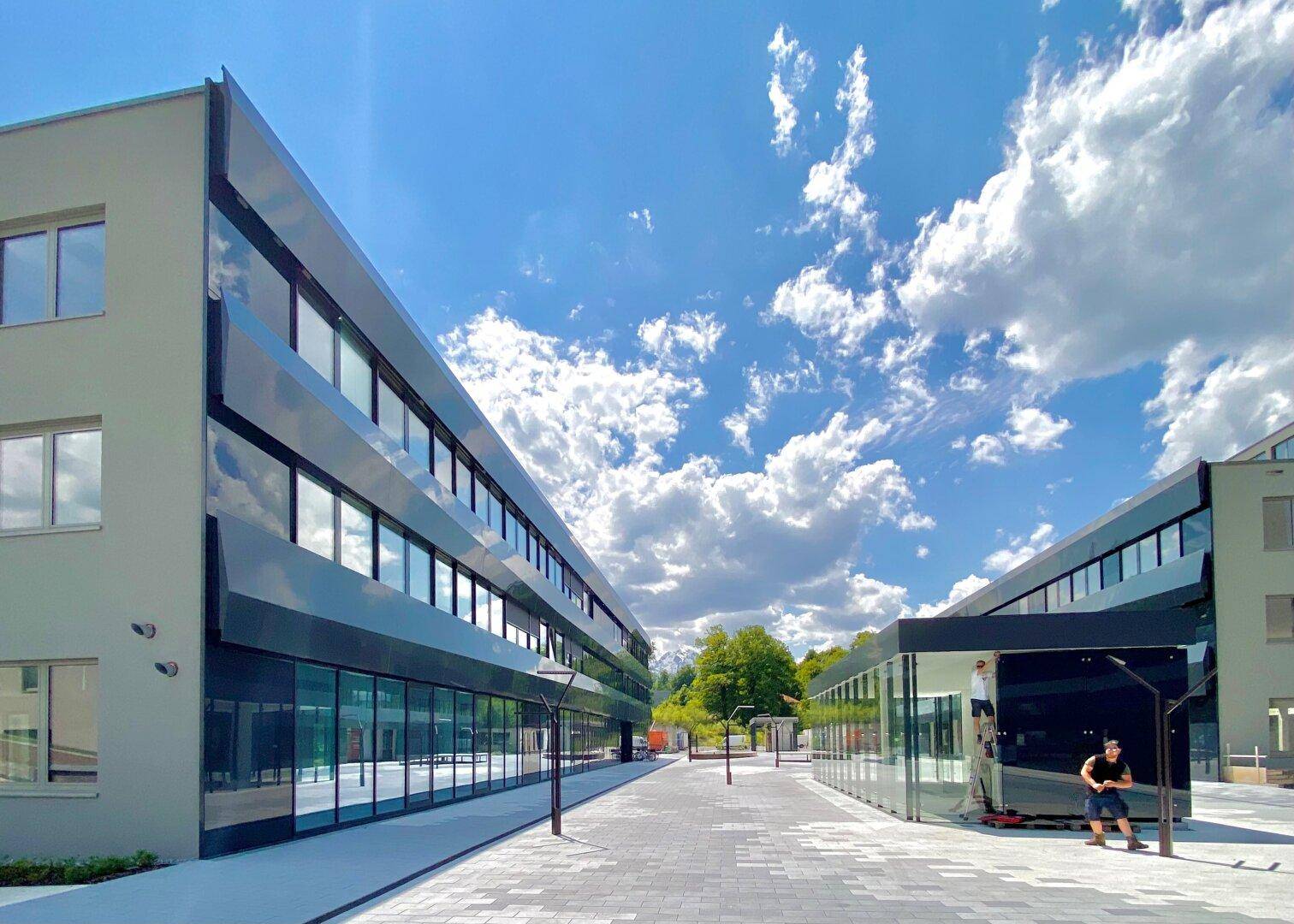 Weitläufiger Außenbereich zwischen modernen Gebäuden mit Glasfassaden und gepflastertem Boden unter strahlendem Himmel.