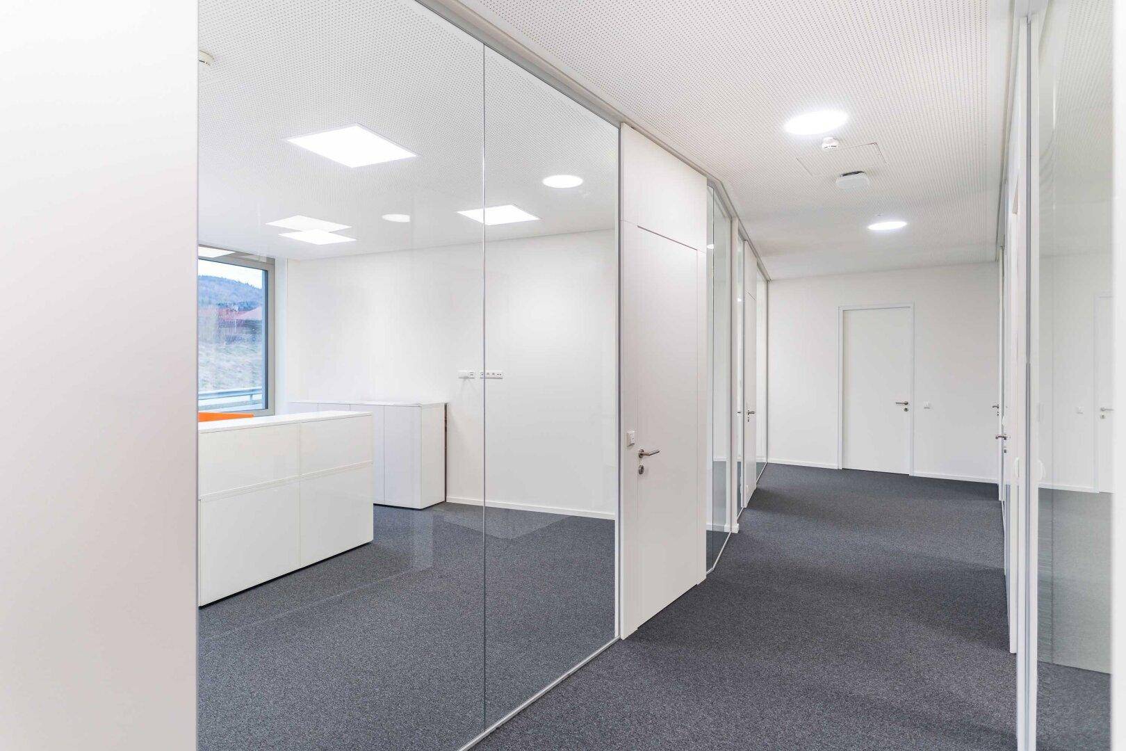 Moderner Bürobereich mit Glaswänden, weißen Schränken und grauem Teppichboden, hell und funktional.