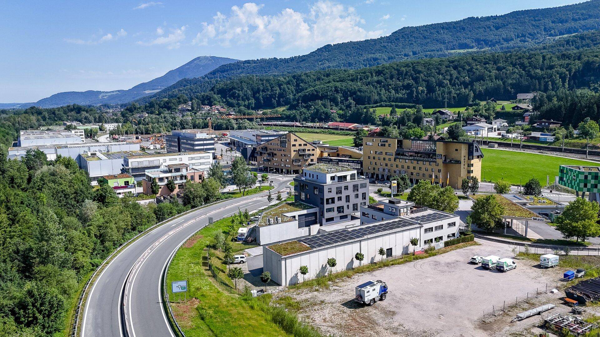 Weitläufige Luftaufnahme eines Gewerbegebiets mit modernen Gebäuden und grüner Umgebung vor Bergkulisse.