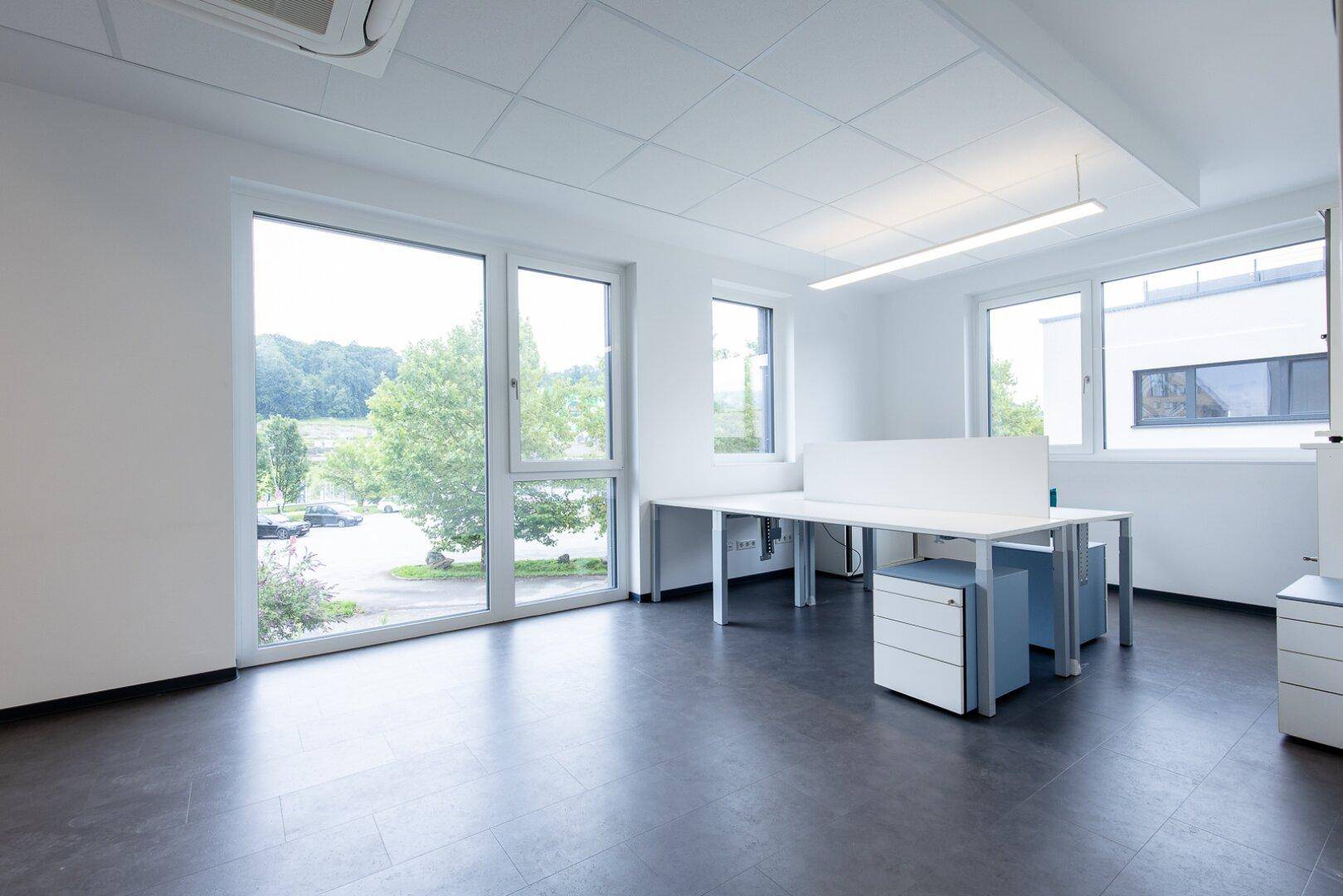 Helles und modernes Büro mit großen Fenstern, die viel Tageslicht hereinlassen und eine grüne Aussicht bieten.