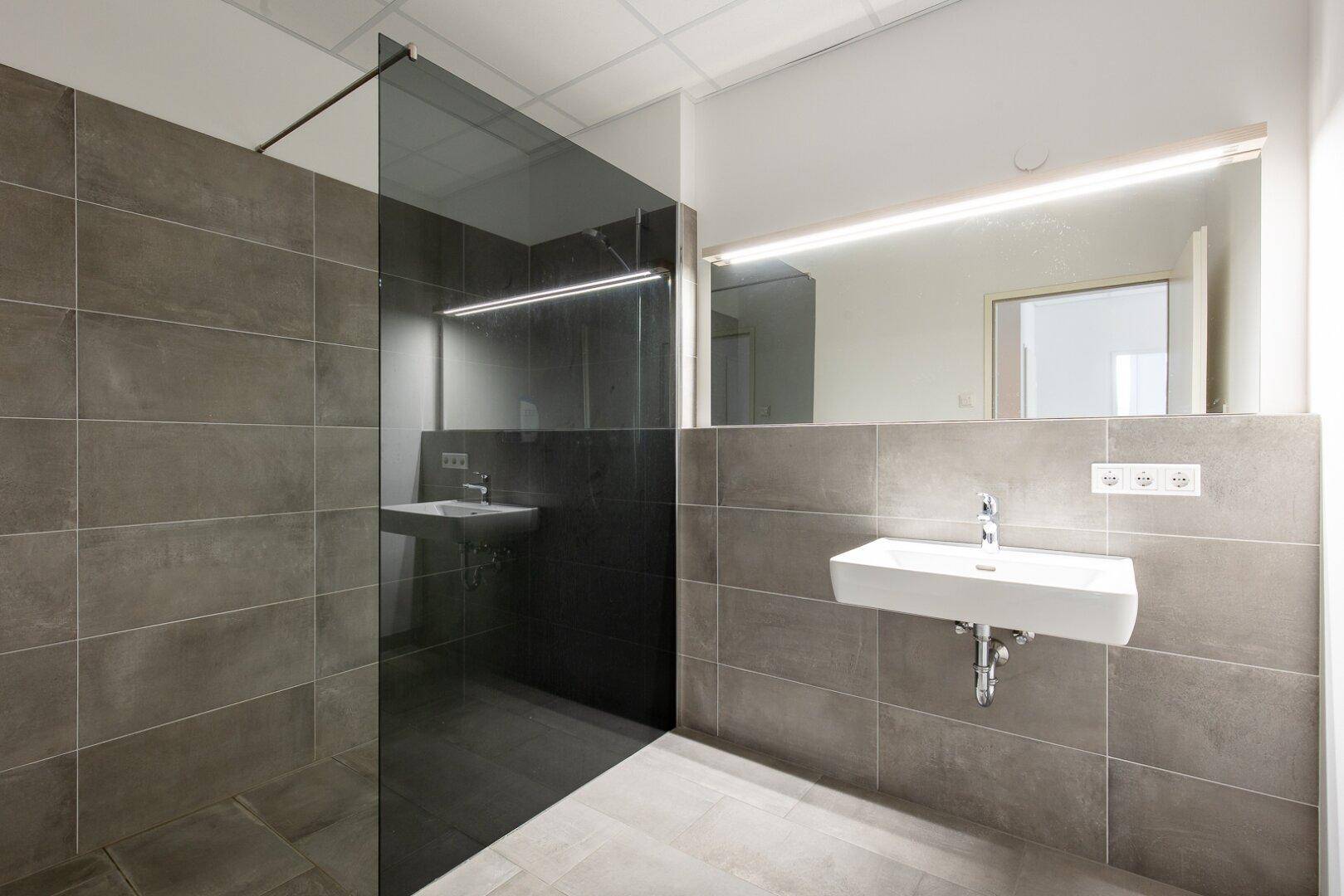 Modernes Badezimmer mit grauen Fliesen, zwei Waschbecken und einer geräumigen begehbaren Dusche mit Glaswand.