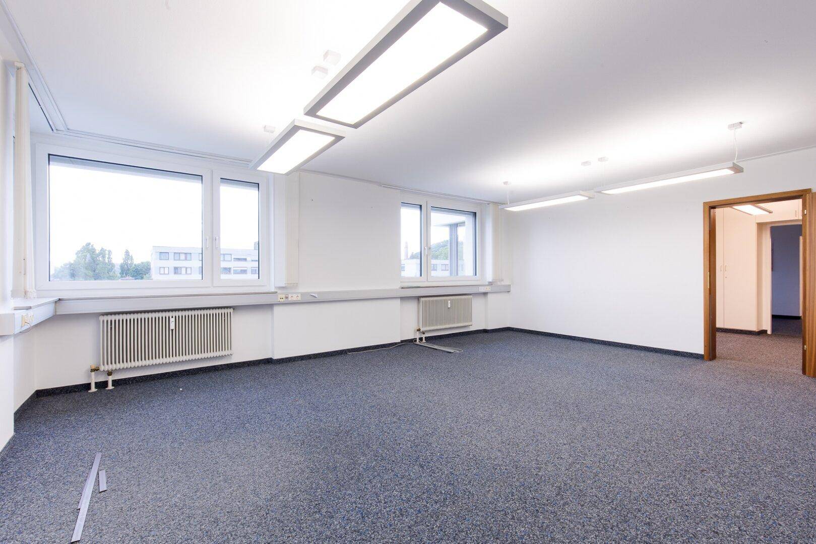 Geräumiges, leeres Büro mit Teppichboden, großen Fenstern und Deckenbeleuchtung, bereit zur Einrichtung.