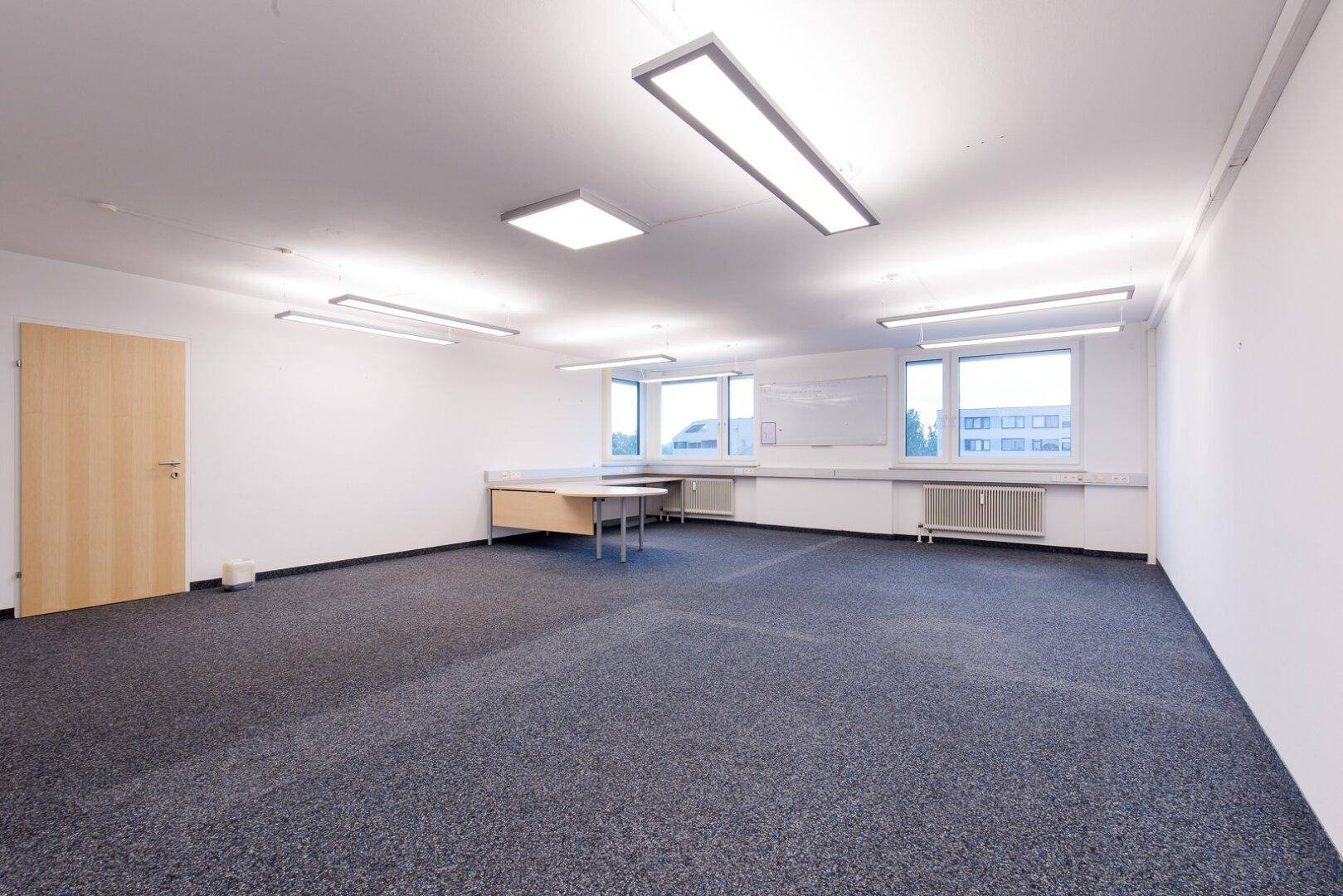Geräumiges, leeres Büro mit Teppichboden, großen Fenstern und heller Deckenbeleuchtung, bereit zur individuellen Gestaltung.