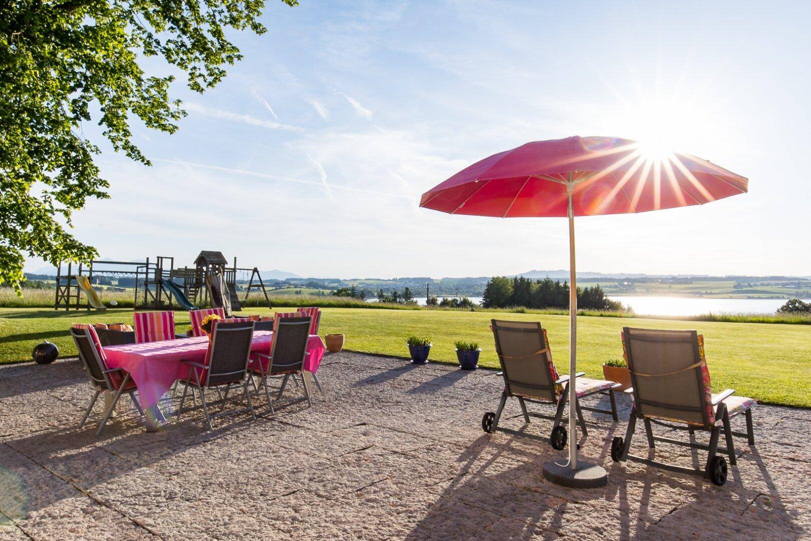 Sonnige Terrasse mit Esstisch, Liegestühlen und Blick auf den See und Spielplatz.
