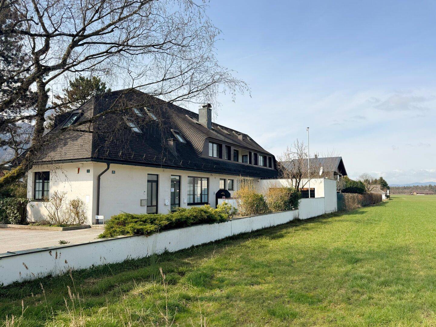 Großes Gebäude mit dunklem Dach und weißer Fassade, umgeben von einer grünen Wiese unter blauem Himmel.