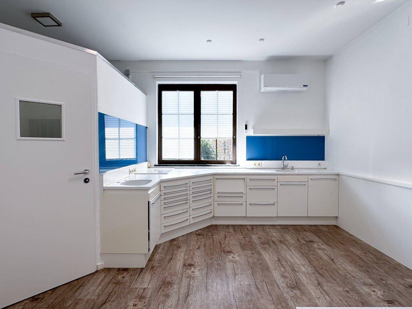 Funktionale Küchenzeile mit weißen Schränken, blauer Rückwand und Fenster, ideal für eine Büroumgebung.
