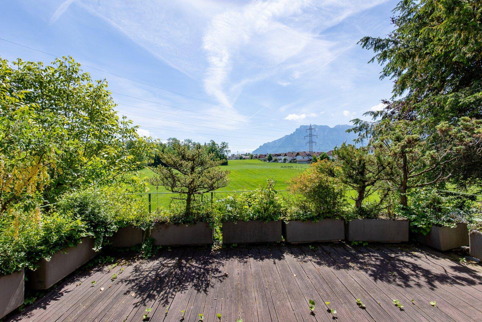 Geräumige Holzterrasse mit Pflanzkübeln und weitem Blick über grüne Felder und Berge.