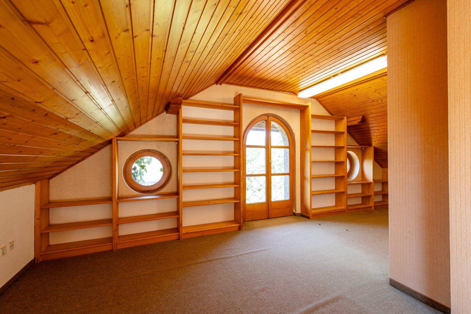 Gemütlicher Dachbodenraum mit Holzverkleidung, eingebauten Regalen und einem runden Fenster.