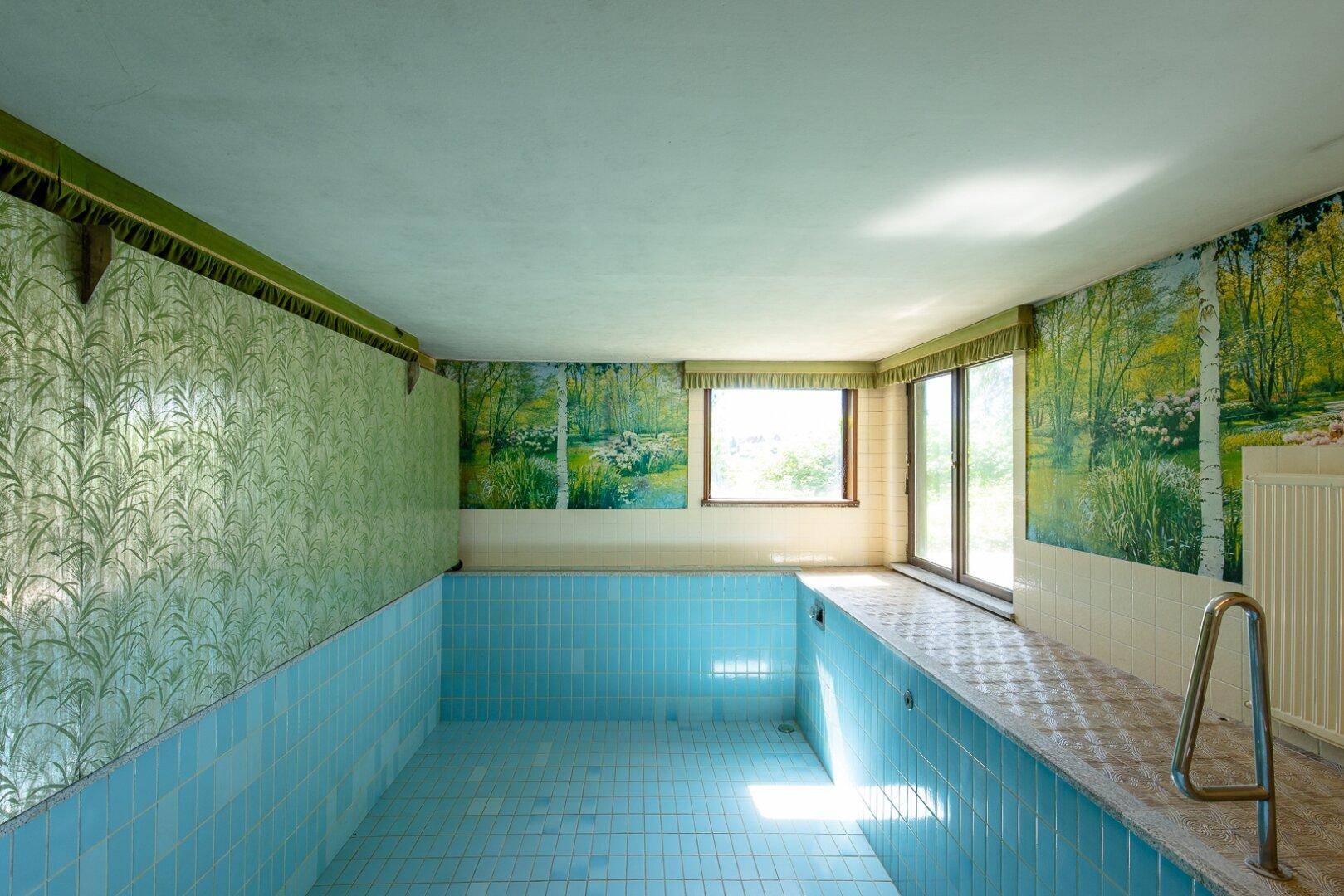 Innenpoolbereich mit blauen Fliesen und grüner Tapete, der eine Renovierung benötigt.