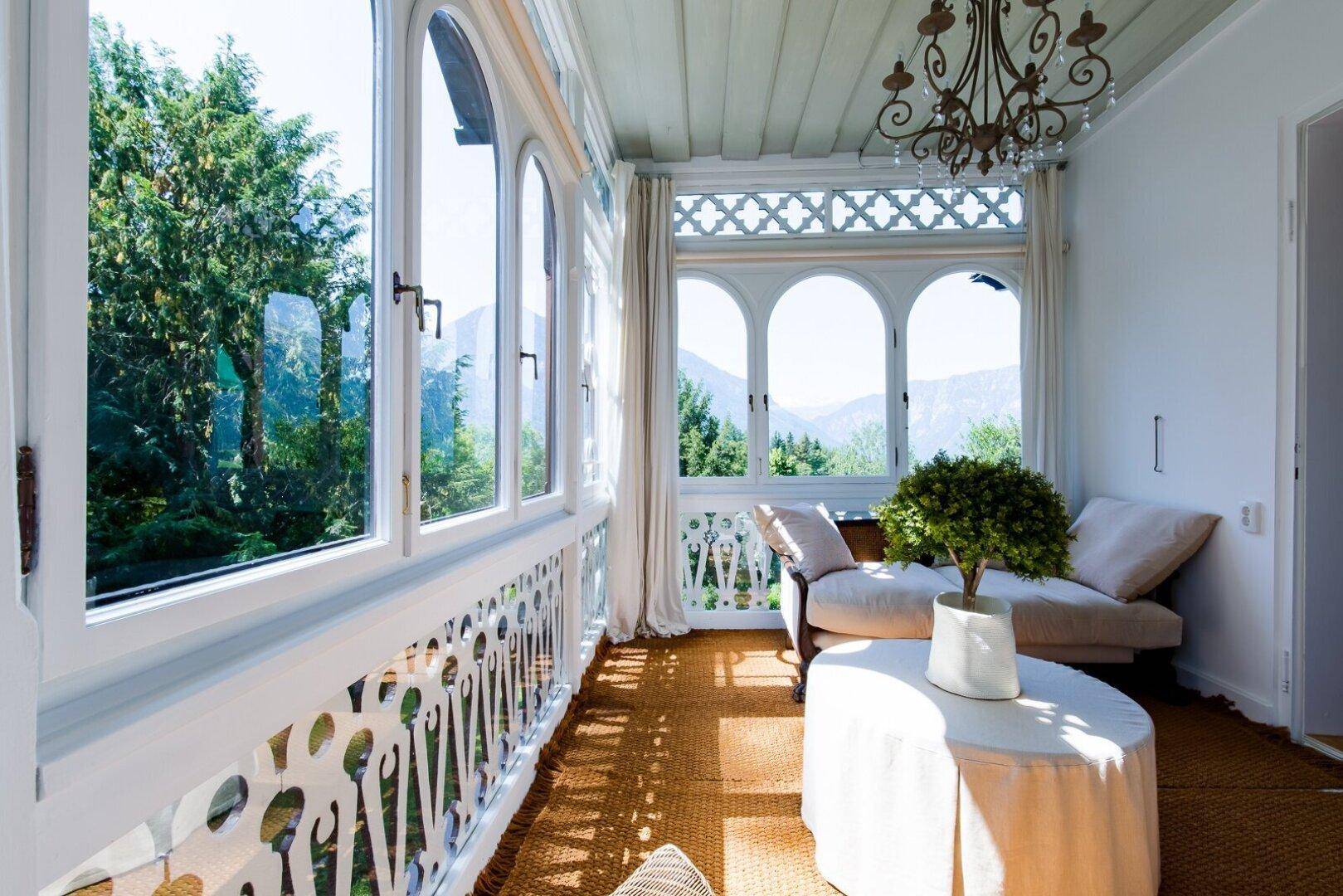 Heller Wintergarten mit großen Bogenfenstern, gemütlicher Einrichtung und Panoramablick auf die Berge.