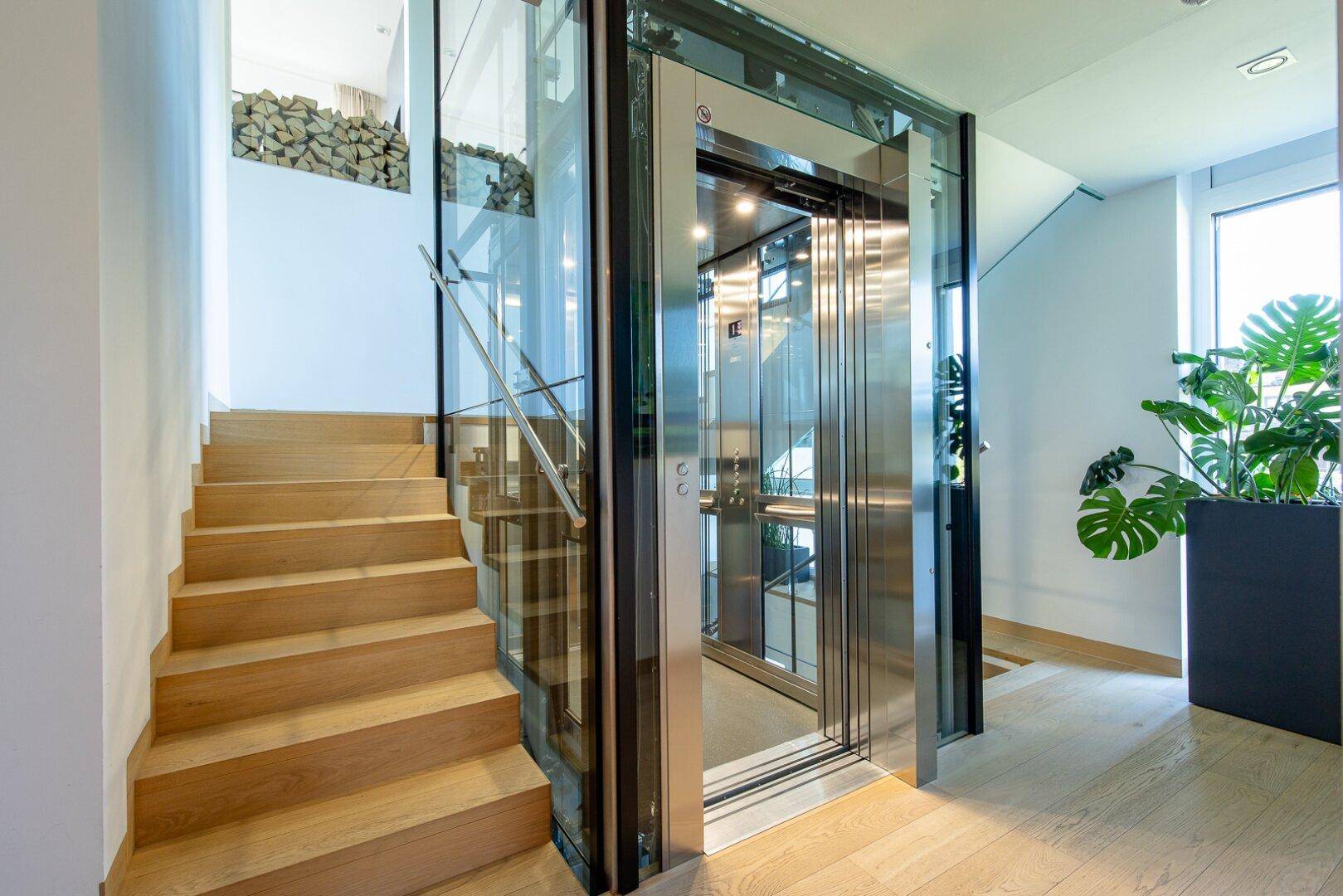 Heller Eingangsbereich mit moderner Holztreppe und einem eleganten Glasaufzug.