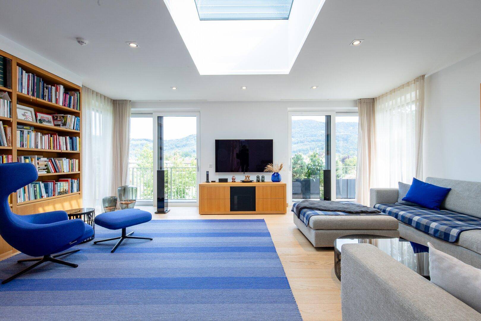 Helles Wohnzimmer mit großem Bücherregal, blauem Teppich und Zugang zum Balkon mit Bergblick.