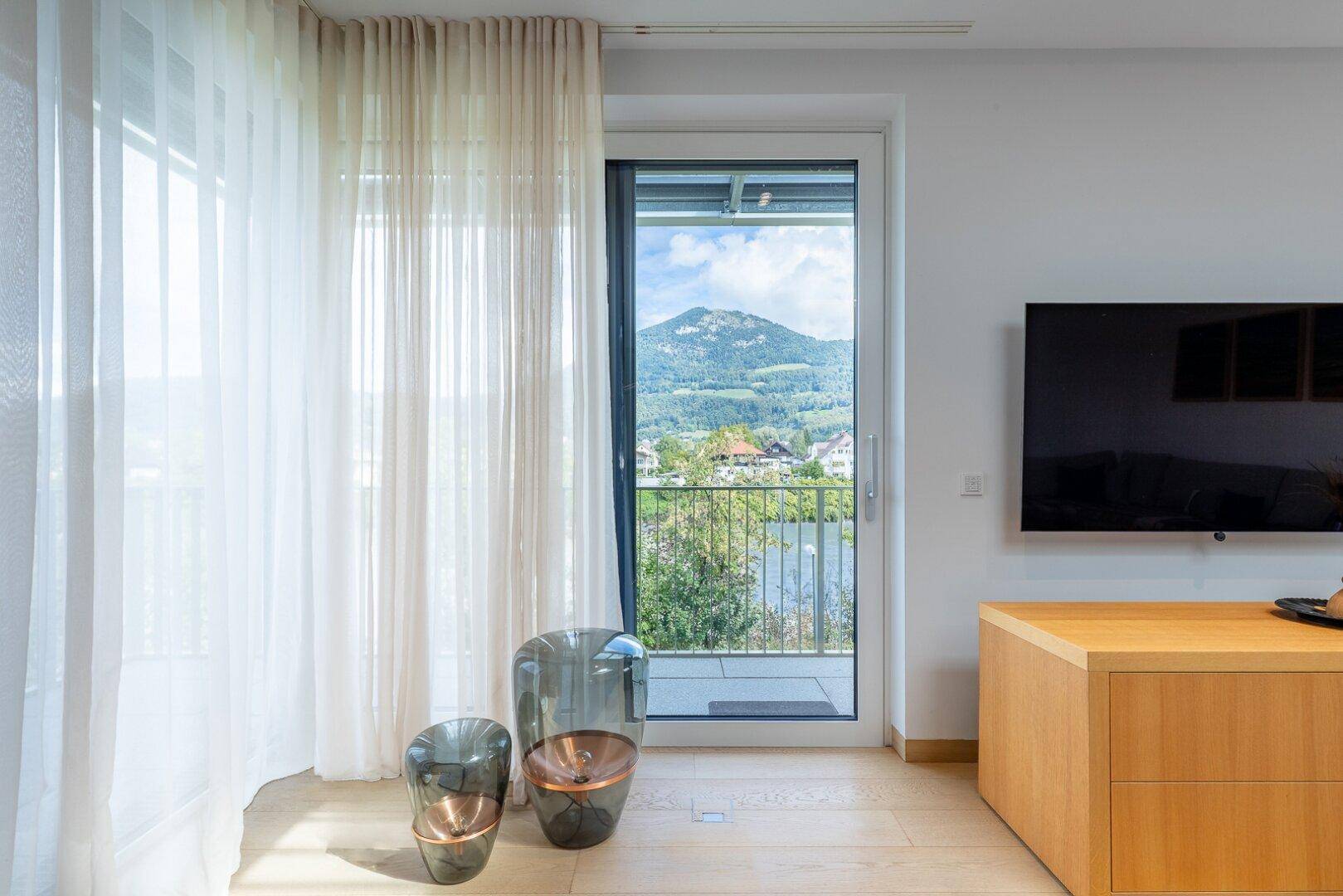 Große Fensterfront im Wohnzimmer mit Blick auf die Berge und den See, umgeben von hellen Vorhängen.