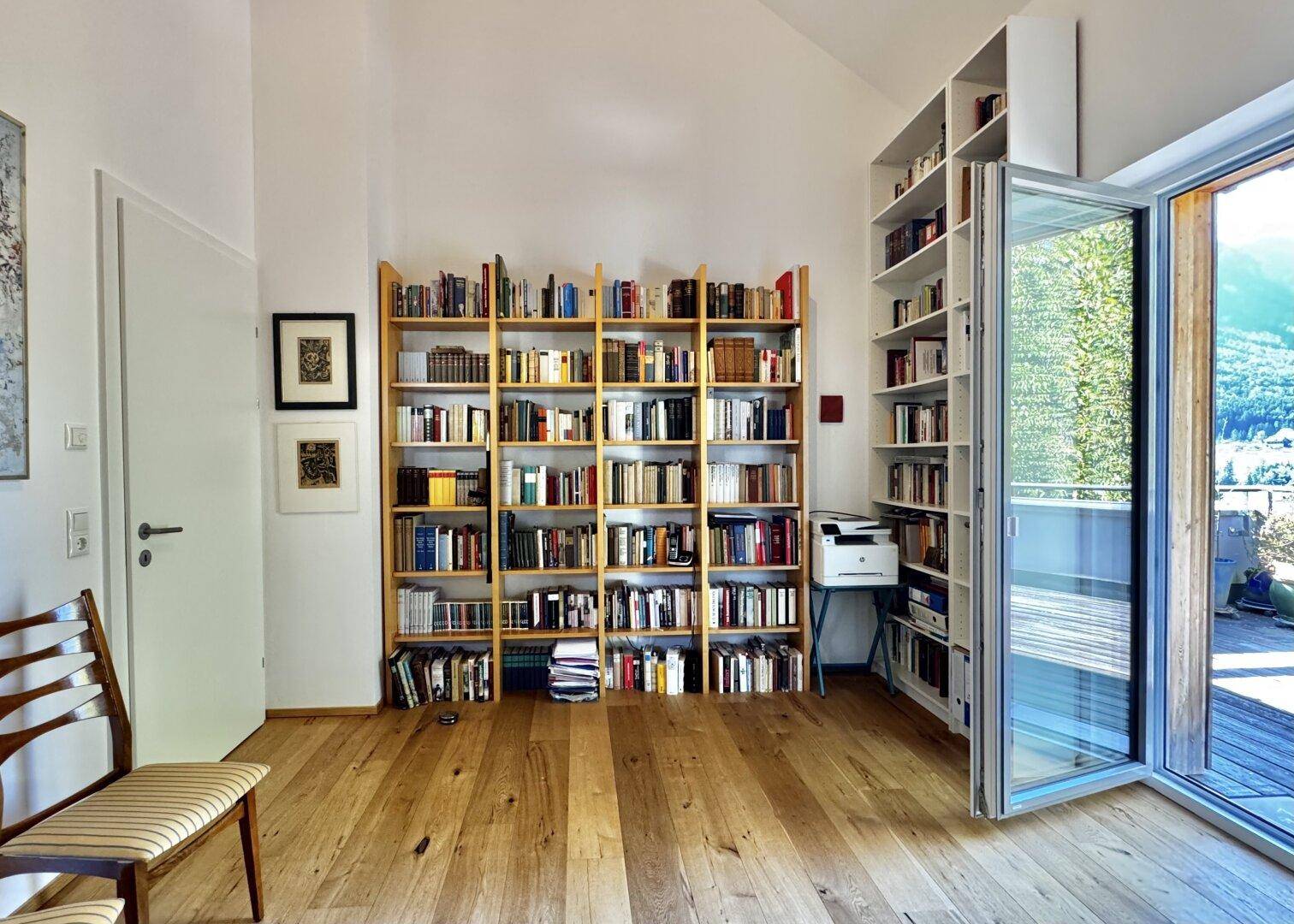 Lichtdurchfluteter Raum mit Holzboden, Bücherregalen und Zugang zu einer Terrasse mit Bergblick.