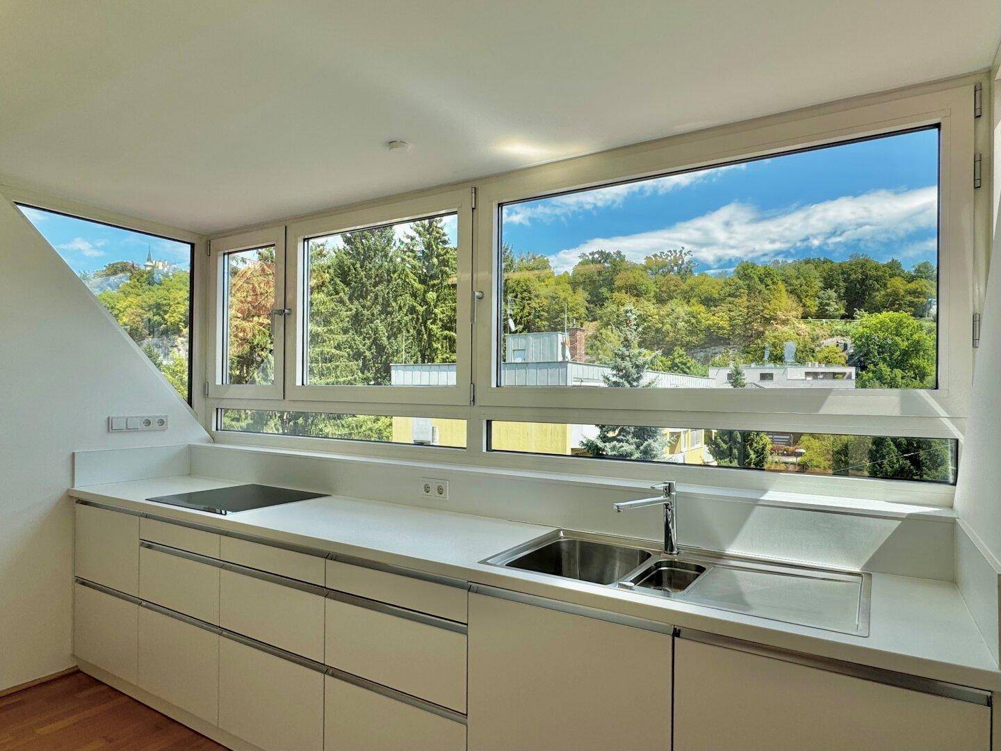 Moderne Küche mit weißen Schränken, Arbeitsplatte und breiten Fenstern mit Grünblick.