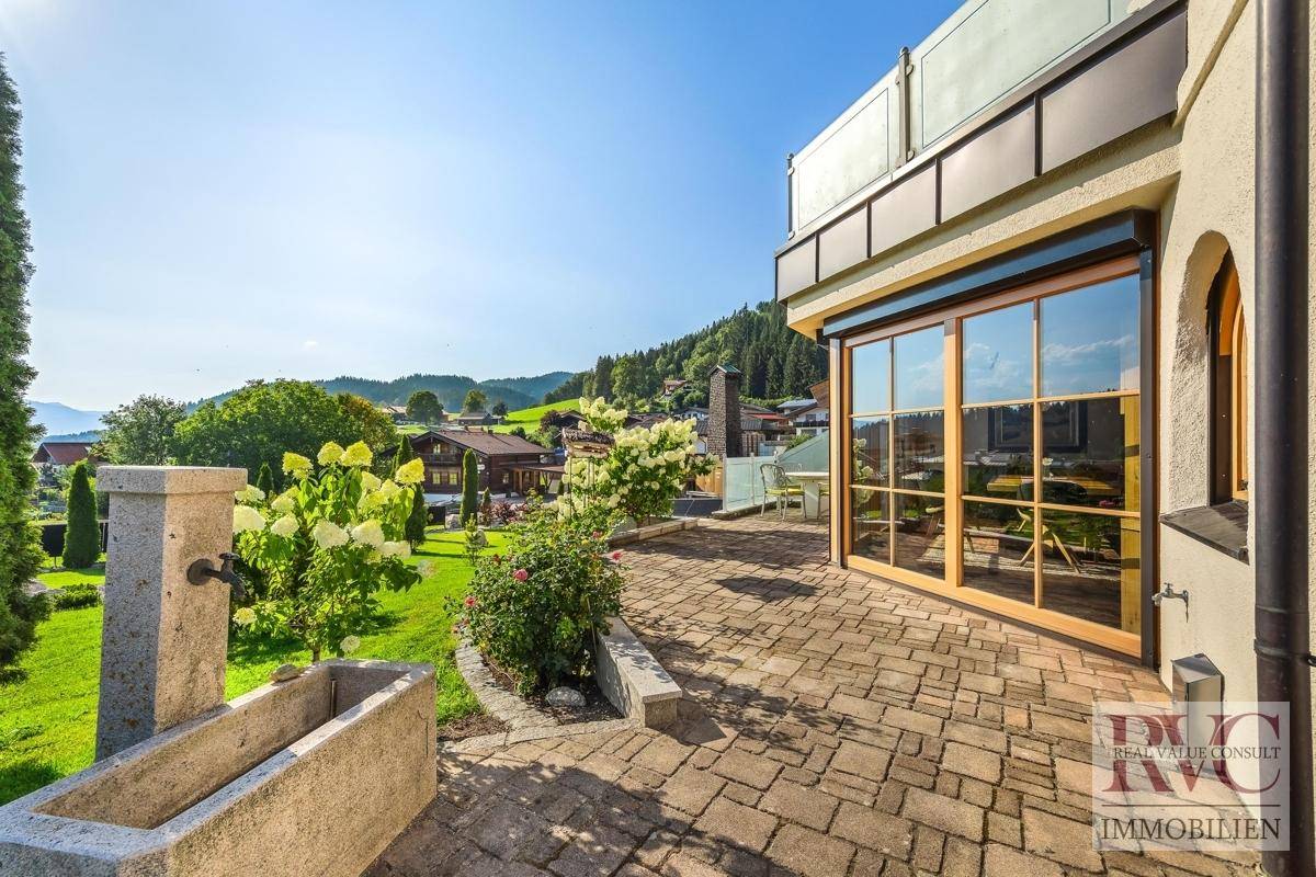 Gepflasterte Terrasse mit Wasserspiel, Blumenbeeten und Zugang zum grünen Gartenbereich.