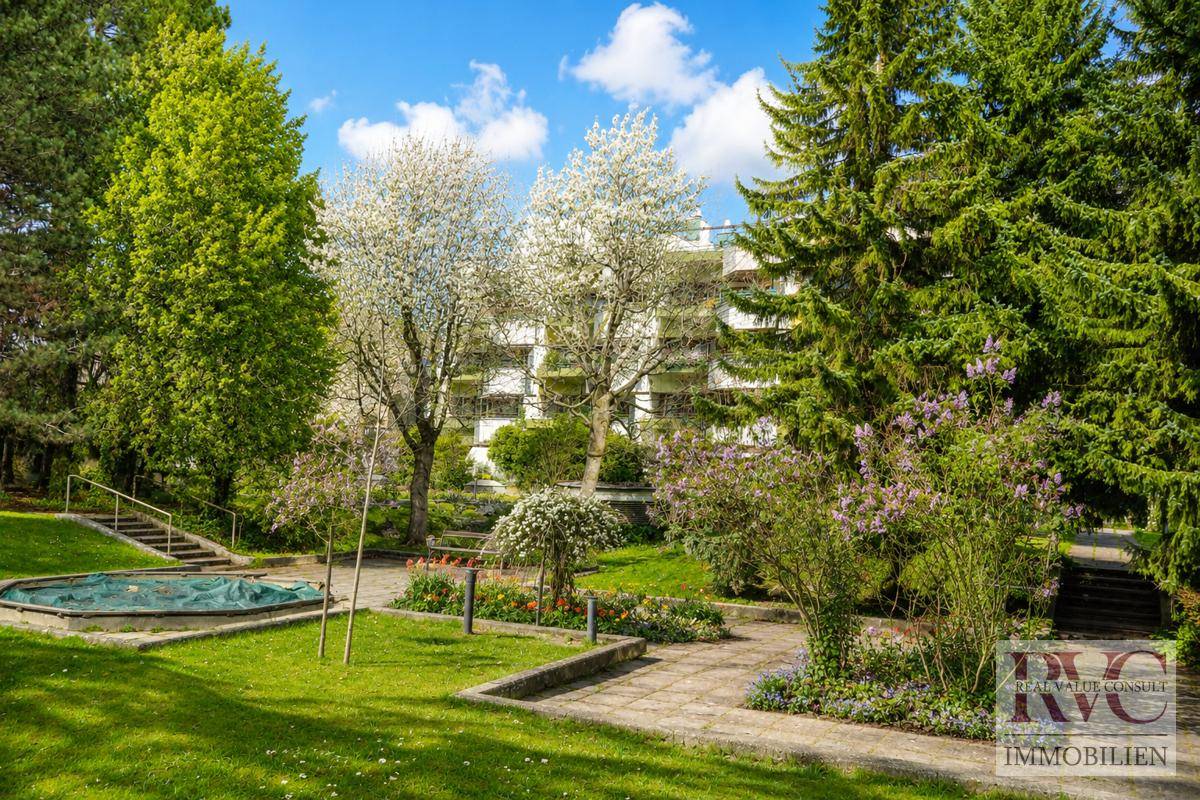Weitläufige Gartenanlage mit Bäumen, Sträuchern und einem kleinen Teich vor dem Gebäude.