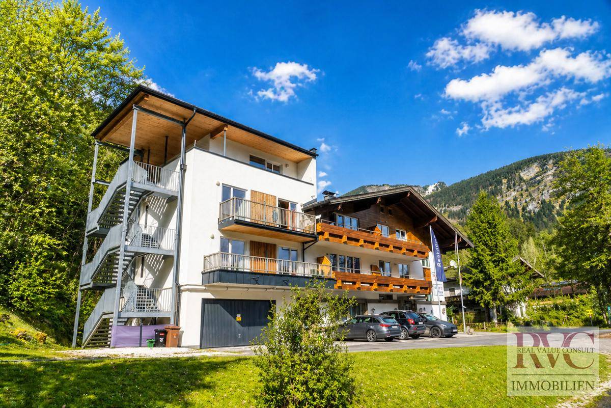 Modernes Gebäude mit traditionellen alpinen Elementen in grüner Berglandschaft unter blauem Himmel.
