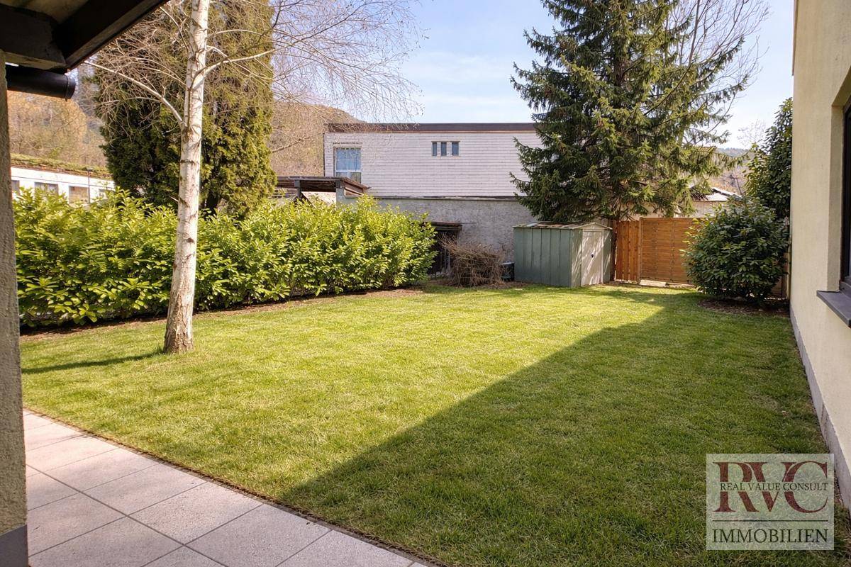 Sonniger Gartenbereich mit Rasenfläche, Hecken und einem Gartenhaus im Hintergrund.