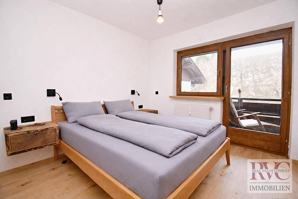 Schlafzimmer mit Holzbett, großen Fenstern und Zugang zu einem Balkon.