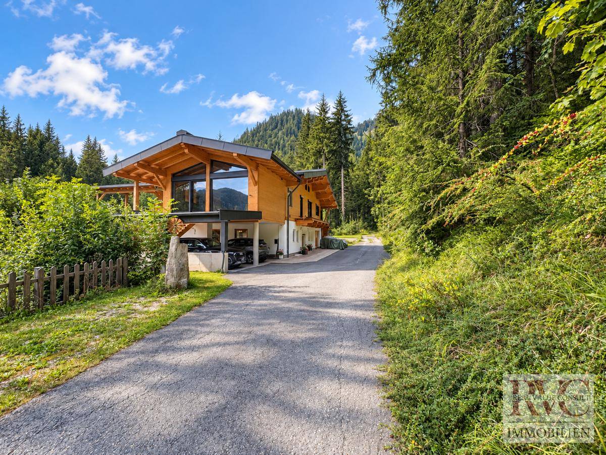 Zufahrt zum modernen Holzhaus mit Carport, umgeben von dichtem Wald und alpiner Landschaft.