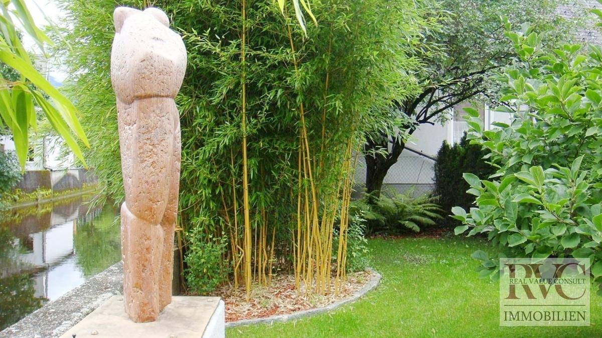 Grüner Gartenbereich am Wasser mit Bambuspflanzen und einer modernen Skulptur.