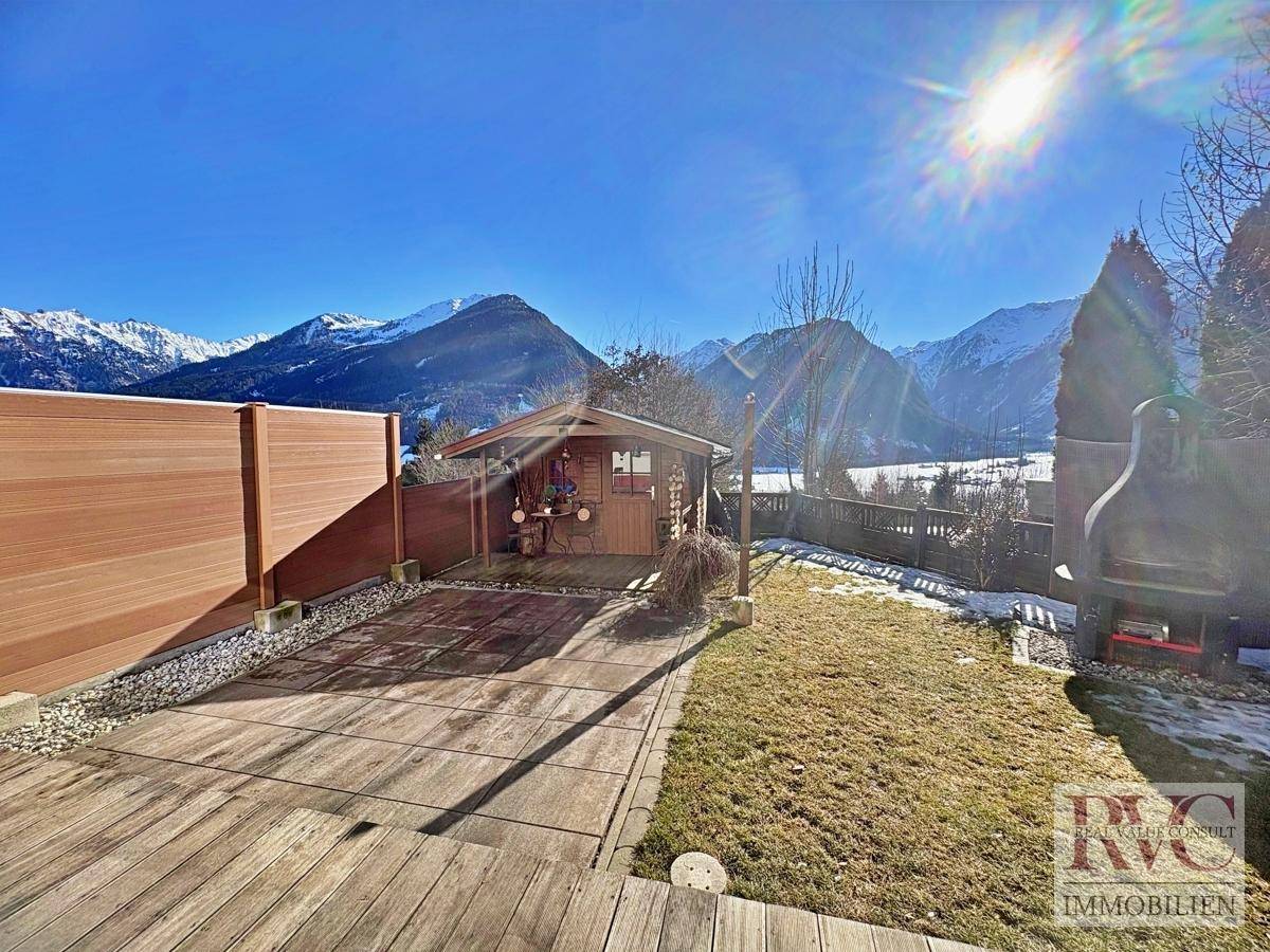 Sonniger Gartenbereich mit Holzterrasse, Gartenhaus und weitem Blick auf die Berge.