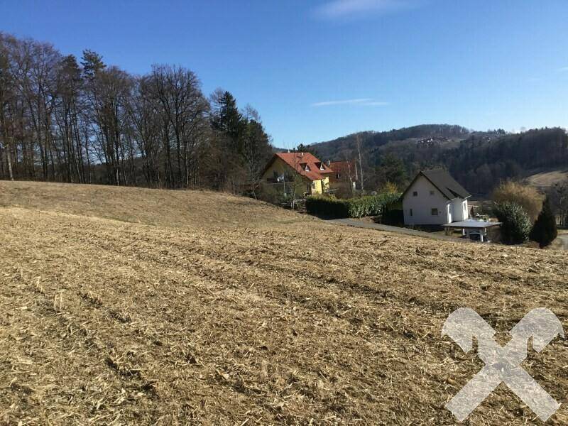 Blick über ein Feld auf mehrere Wohnhäuser in einer ländlichen Umgebung mit Hügeln im Hintergrund.