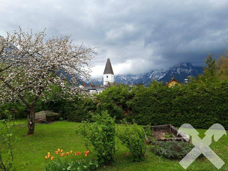Idyllischer Garten mit blühendem Baum, Gemüsebeet und Blick auf Kirche und Berge.