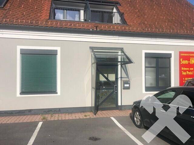 Vorderansicht des Gebäudes mit Eingangsbereich, Fenstern mit Rollläden und einem geparkten Auto davor.