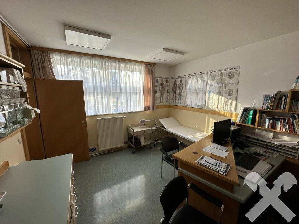 Sprechzimmer mit Schreibtisch, Untersuchungsliege und medizinischen Postern an den Wänden.
