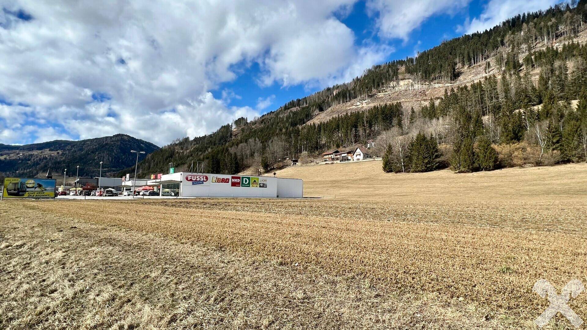 Panoramablick auf ein weites Feld, eine Straße und ein Geschäftshaus vor einer bewaldeten Bergkette unter einem bewölkten Himmel.