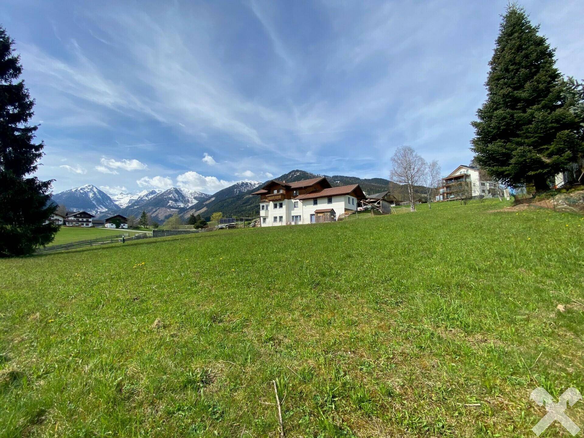 Weitläufige grüne Wiese mit einem Haus im Hintergrund und schneebedeckten Bergen unter blauem Himmel.