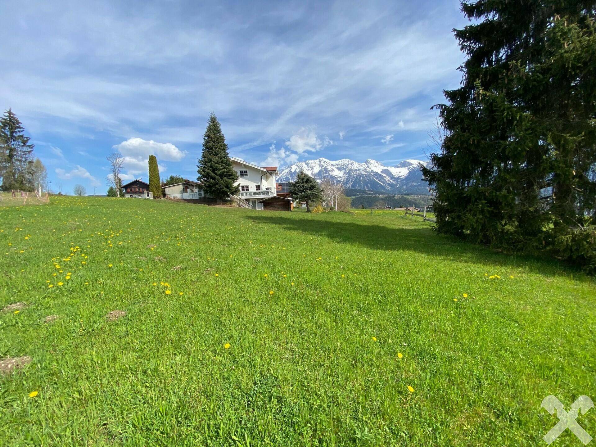 Sonniger Blick über eine grüne Wiese auf ein Haus und eine beeindruckende Kette schneebedeckter Berge.