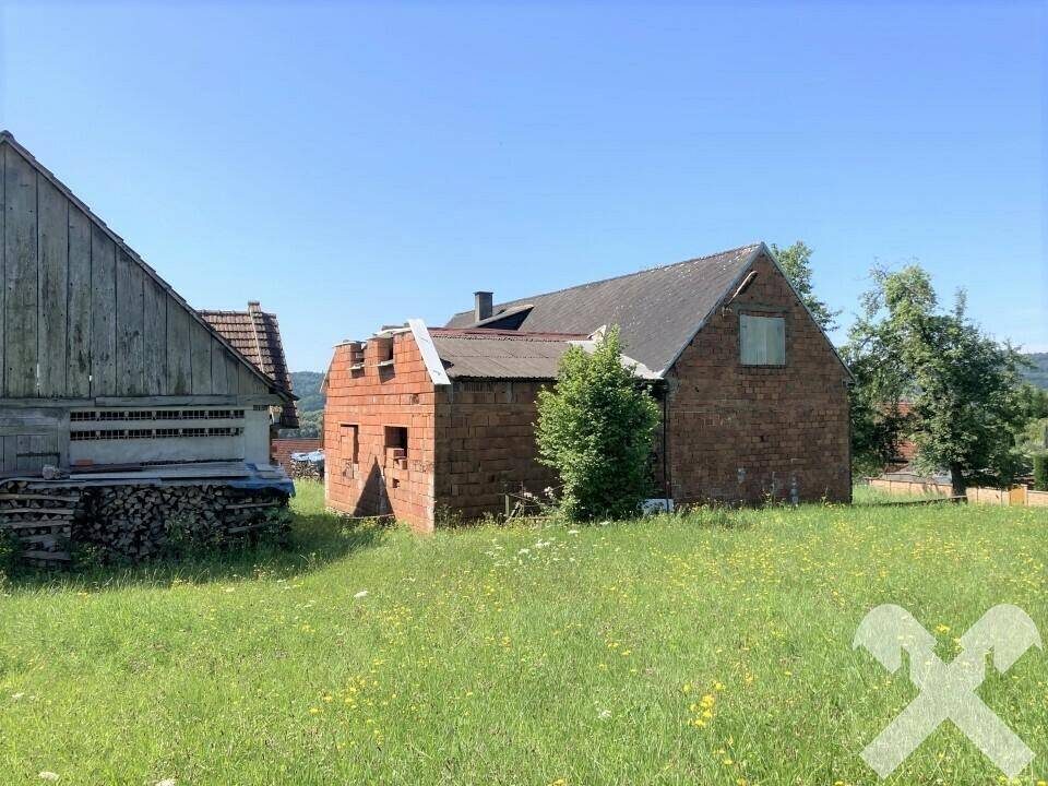 Ländliche Ansicht mit einem alten Holzgebäude und einem Backsteingebäude im Bau, umgeben von einer grünen Wiese unter klarem Himmel.