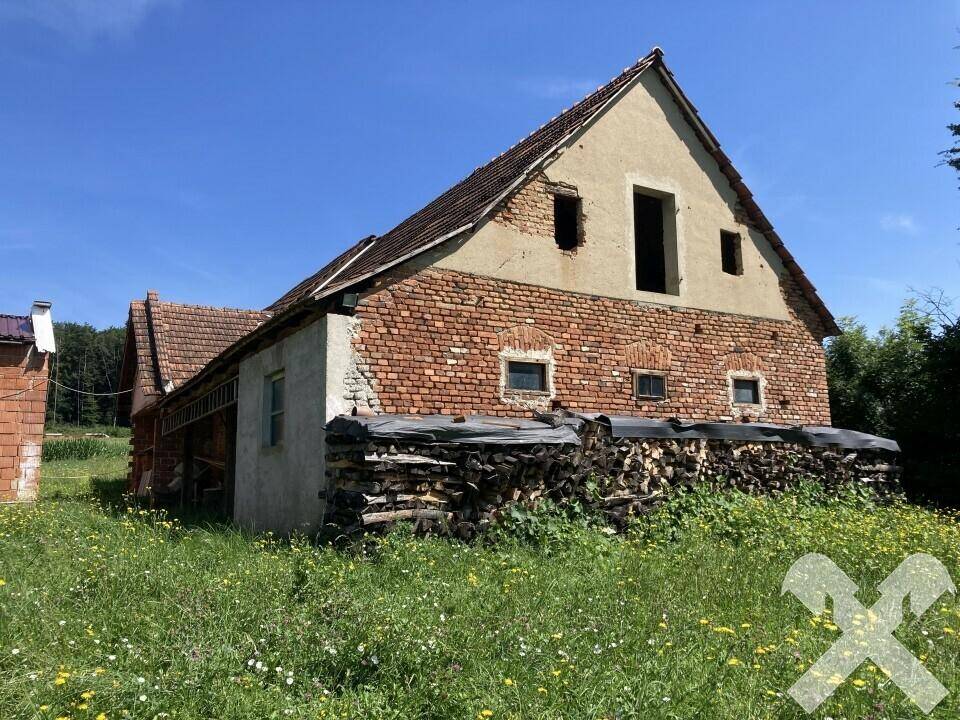 Seitenansicht eines älteren Backsteingebäudes mit Ziegeldach und Holzstapeln davor auf einer Wiese.