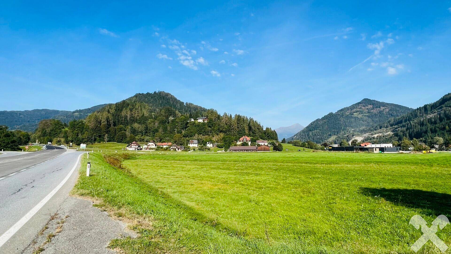 Panoramablick über eine weite grüne Landschaft mit Bergen, einer Straße und vereinzelten Häusern am Horizont.