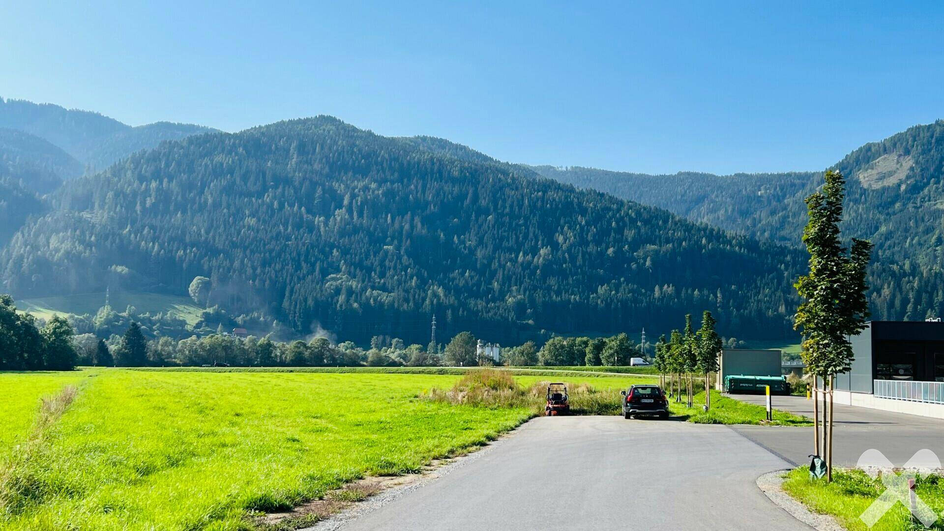 Blick auf eine grüne Wiese mit bewaldeten Bergen, einer Zufahrt und einem Gebäude im rechten Bildbereich.